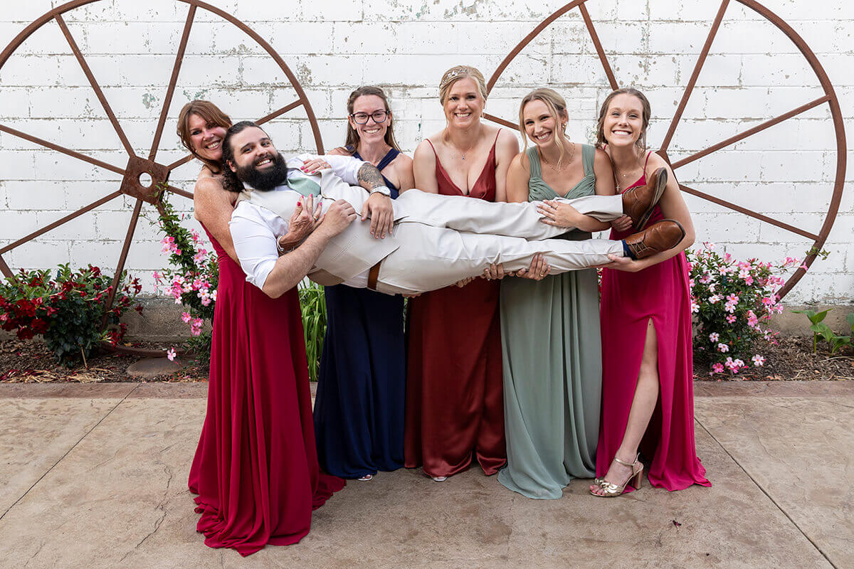 Five women dressed elegantly holding a man in the air wearing a tuxedo, celebrating fun and style for formal occasions